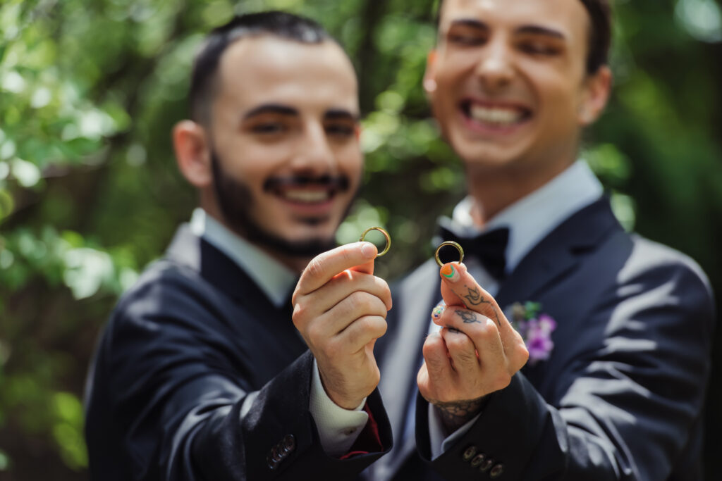 Humblebee Wedding Films Destination Wedding Videographer, happy and blurred gay men holding wedding rings in hands.