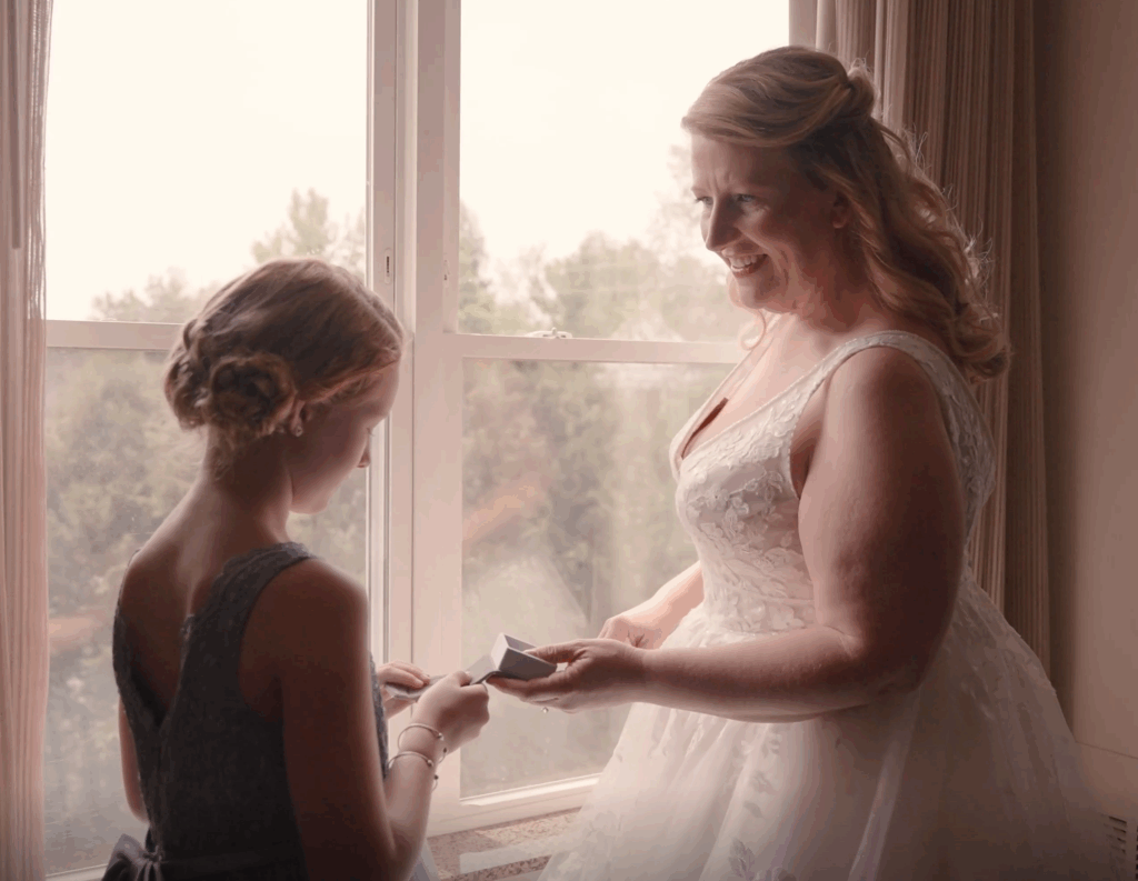 Humblebee Wedding Films Birch Wood Vineyards, a flower girl and bride stand before a window discussing the wedding bands