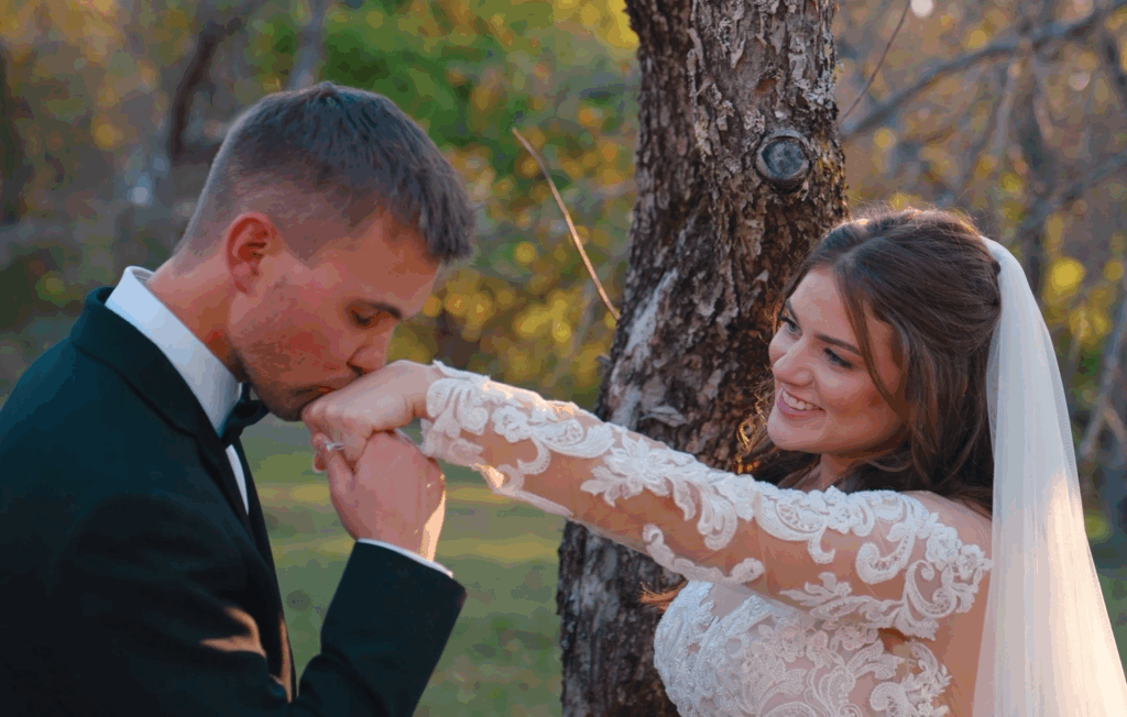 Humblebee Wedding Films Questions To Ask Wedding Videographer Questions To Ask Photographer & Videographer Newlywed couple poses in front of tree, groom kisses bride's hand