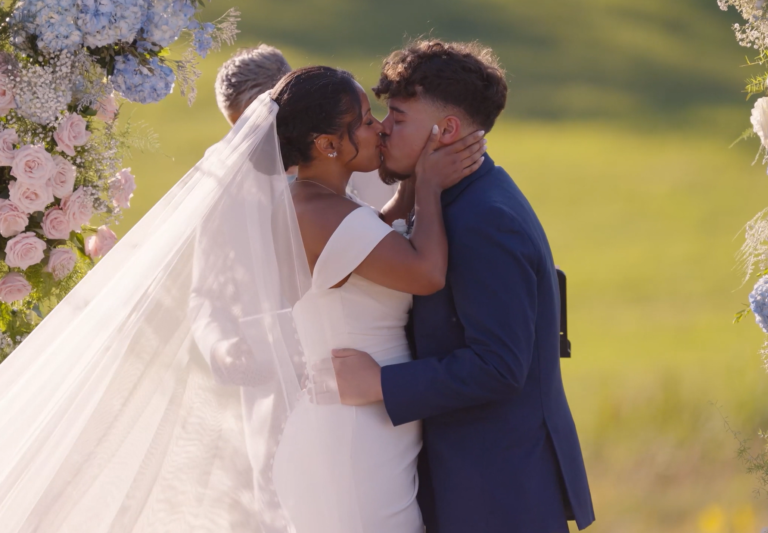 Humblebee Wedding Films Wedding Videographer, newlyweds embrace and share a kiss under a floral arch
