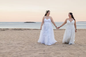 Humblebee Wedding Films Wedding venues nh seacoast, brides walk hand in hand on the beach with the ocean behind them