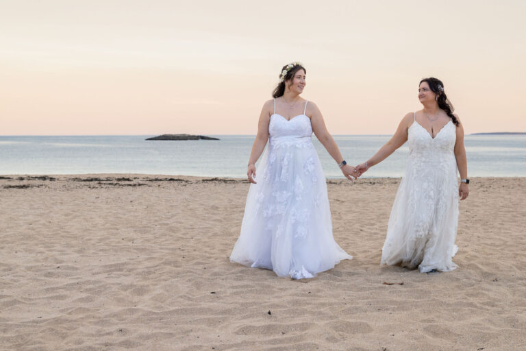 Humblebee Wedding Films Wedding venues nh seacoast, brides walk hand in hand on the beach with the ocean behind them